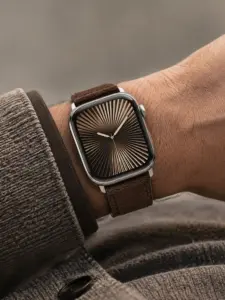 Handgelenk von einem Mann mit Cardigan Ärmel in Braun. Er trägt eine silberne Apple Watch am Handgelenk mit einem dunkelbraunen Wildlederarmband von vild.