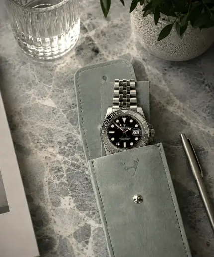 Uhrenetui Leder in Grau mit Rolex GMT Master II Bruce Wayne im Etui und auf Leder Patch liegend. Set Motiv auf Marmortisch mit Buch, Glas Wasser und Pflanze.