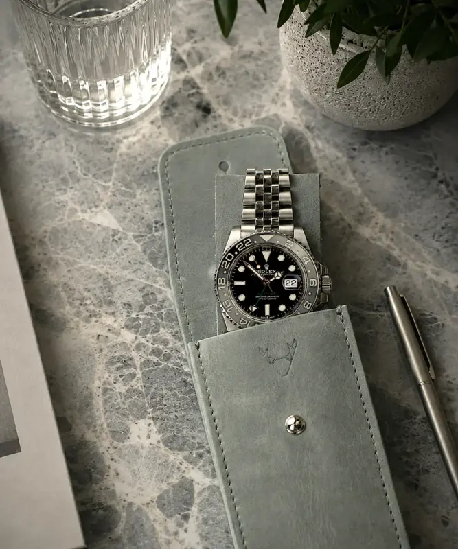 Uhrenetui Leder in Grau mit Rolex GMT Master II Bruce Wayne im Etui und auf Leder Patch liegend. Set Motiv auf Marmortisch mit Buch, Glas Wasser und Pflanze.