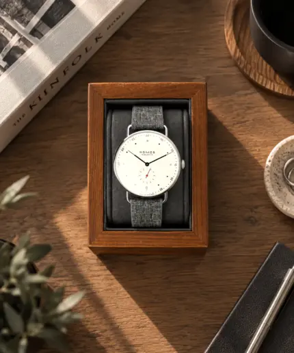 Wooden watch holder in walnut brown and dark grey cushion with Nomos Metro placed on a wooden table with decoration.