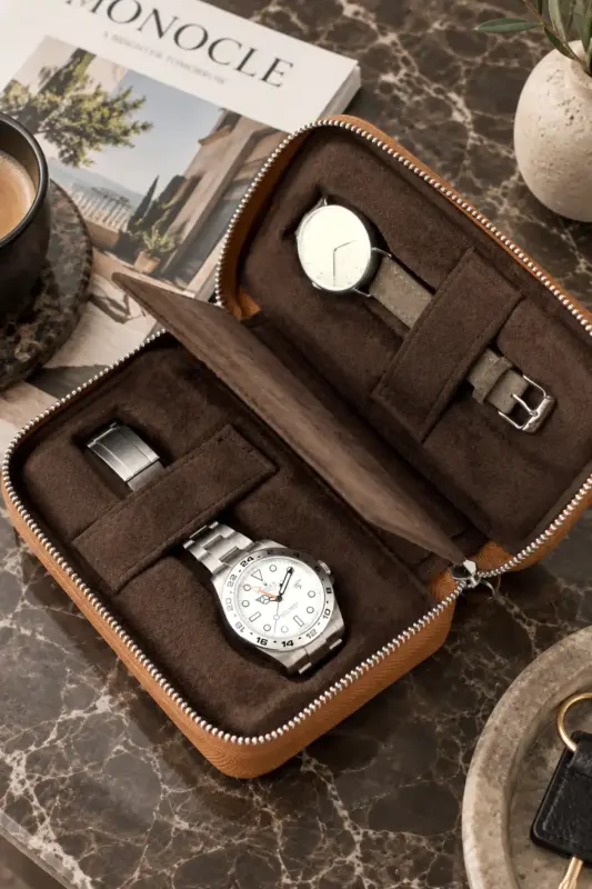An open watch case with a soft, dark brown suede lining. A Rolex Explorer II is clipped into one slot. A Nomos Metro Date is in the second slot. In the background is a stone table with decorative items such as a magazine and a coffee cup.