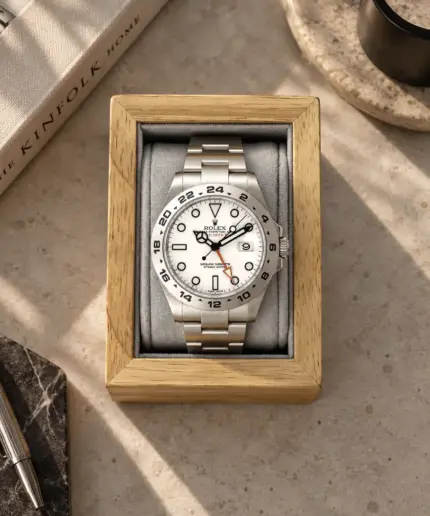 Wooden watch holder in oak brown and light grey cushion with Rolex Explorer II placed on a stone table with decoration.