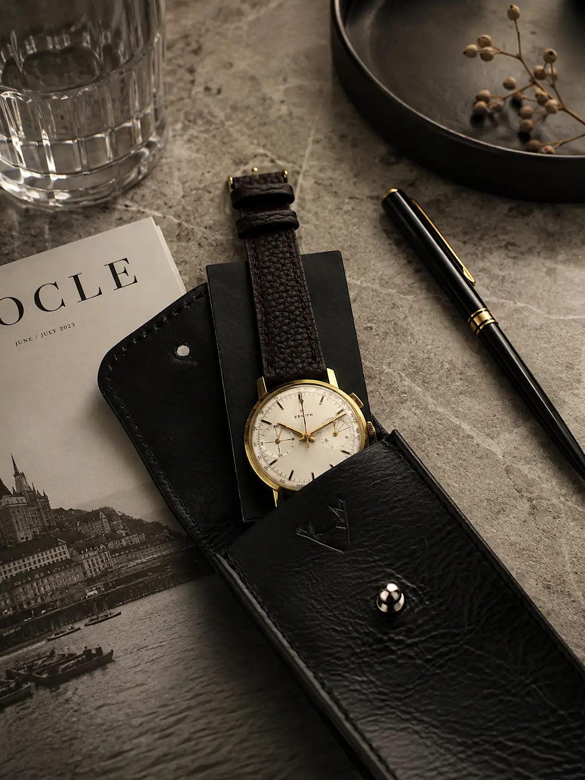 Uhrenetui-Leder-schwarz-1-uhr-motiv-set-zenith-vintage-chrono-gold-vild-hamburg Uhrenetui Leder in Schwarz mit Zenith Chronograph im Etui und auf Leder Patch liegend. Set Motiv auf Tisch mit Magazin, Glas Wasser und Kugelschreiber.