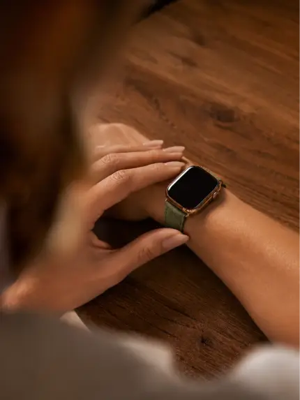 Blick über die Schulter einer Frau auf ihr Handgelenk auf Holztisch mit einer goldenen Apple Watch und grünem Wildlederarmband von vild.