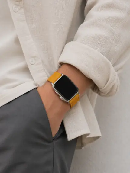 Mann mit Leinen Hemd und grauer Chino steht vor Wand mit Hände in den Hosentaschen und trägt eine silberne Apple Watch mit senffarbenem Lederarmband von vild Hamburg.