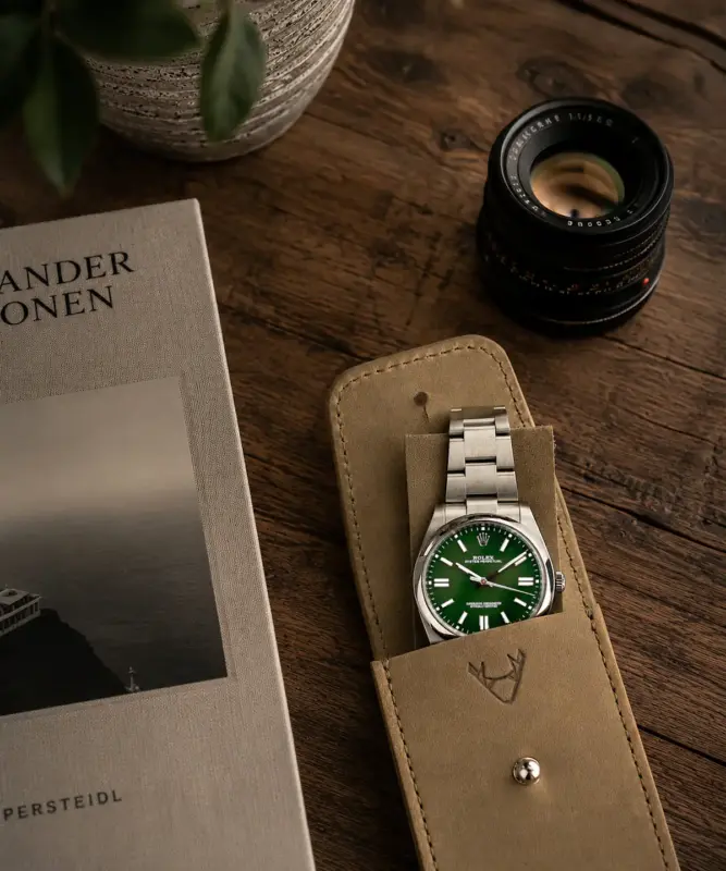 Uhrenetui Leder in Khaki mit Rolex Oyster Perpetual 41mm im Etui und auf Leder Patch liegend. Set Motiv auf Holztisch mit Buch, Kameraobjektiv und Pflanze