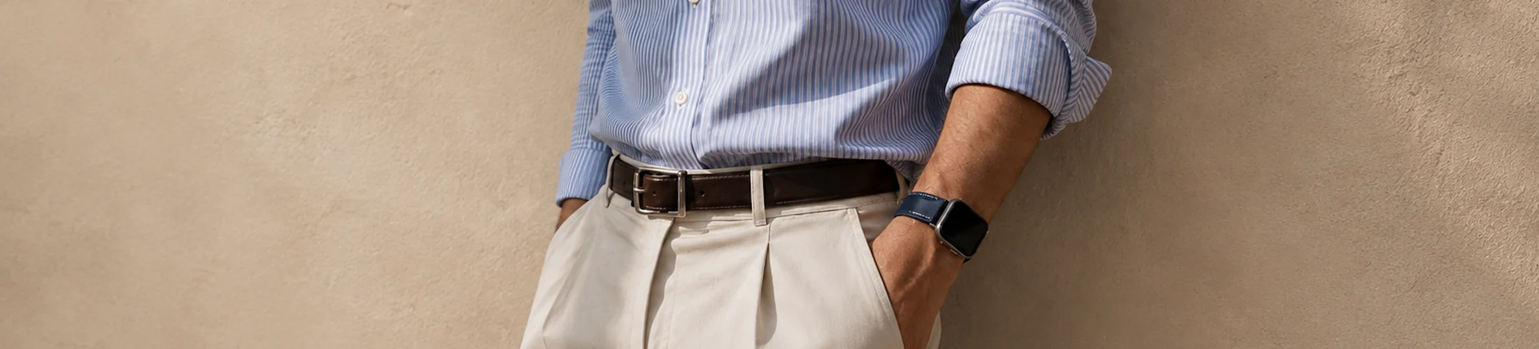 A man in a blue shirt leans against a wall with his hands in his pockets and wears a silver Apple Watch with a dark blue leather strap by vild.