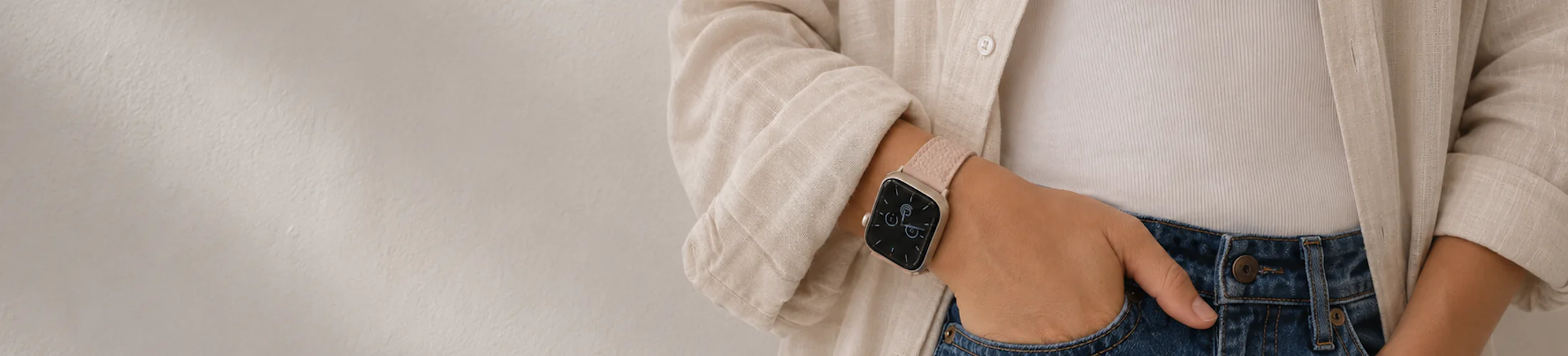 Freu, wearing a linen shirt and jeans, stands in front of a white wall and wears a silver Apple Watch with a pink leather strap.