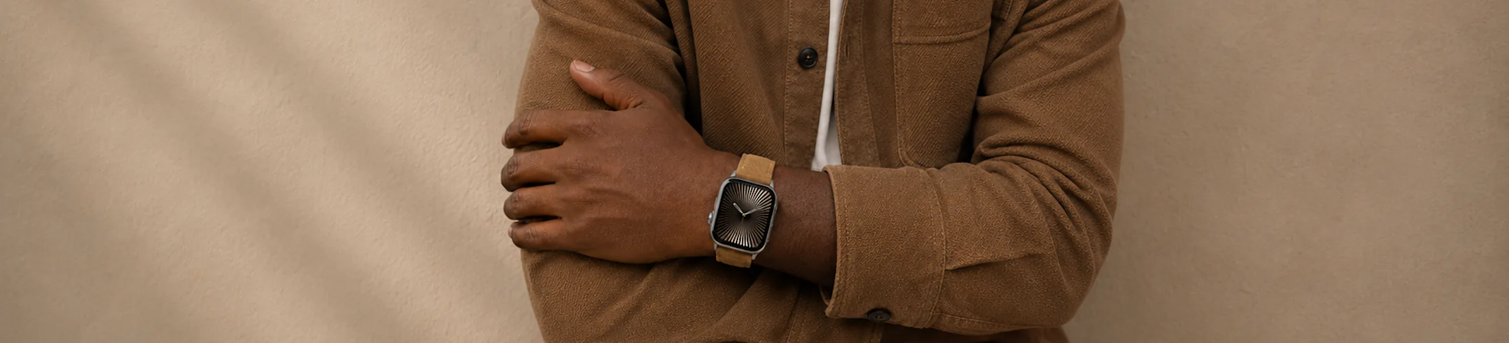 A man wearing a suede shirt stands in front of a wall with his bracelet crossed and wears a silver Apple Watch with a yellow-brown suede strap by vild.