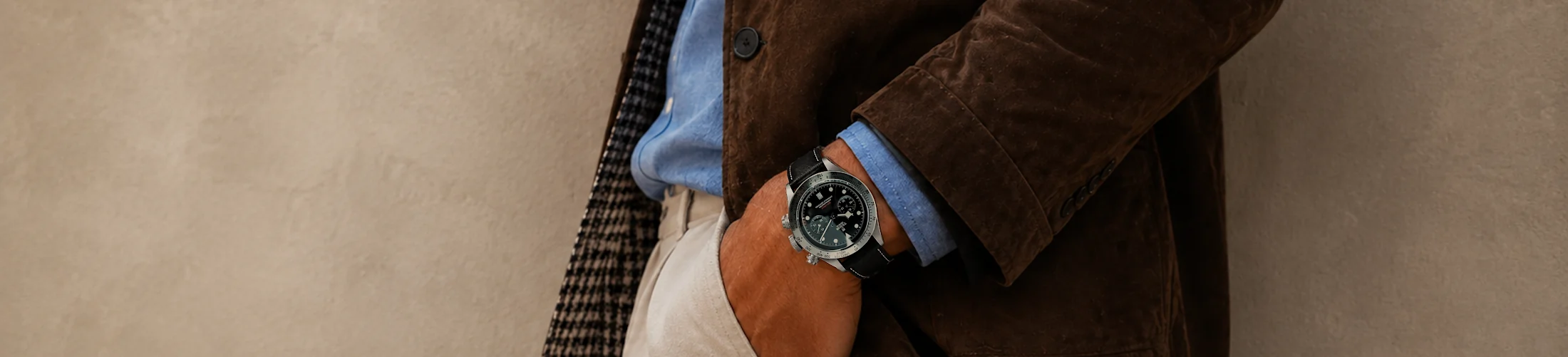 A man in a shirt and jacket, as well as beige chinos, stands in front of a wall with his hands in his pockets and wears a Tudor chronograph with a black leather strap by Vild.