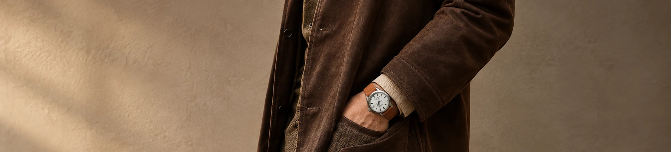 A man in a coat stands with his hands in his pockets in front of a neutral wall and wears a Grand Seiko watch with a light brown suede strap.