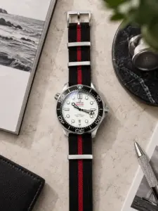 Premium NATO Strap MISTRAL in Schwarz mit rotem Streifen von vild, an einer Omega Seamaster mit weißem Zifferblatt montiert und von oben mit passender Tischdekoration gezeigt