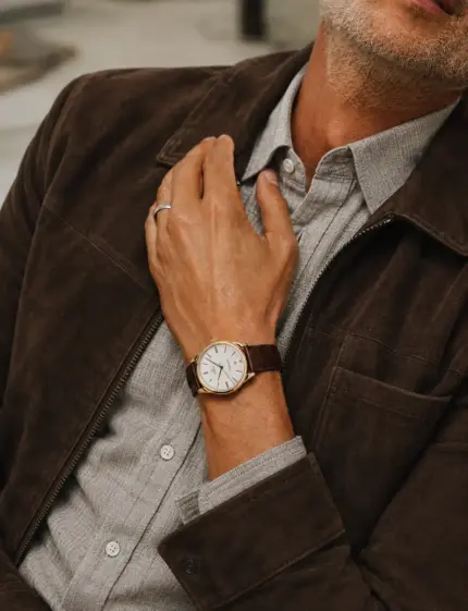 Brown leather crocodile watch strap by vild Hamburg on an Omega De Ville Tresor ref 432.53.40.21.02.002 on the wrist of a man wearing a dark brown nubuck leather jacket and a grey linen shirt.