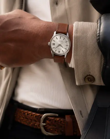 Brown leather watch strap from vild Hamburg on a King Seiko 4402-8000 on the wrist of a man wearing a light cardigan, white knit shirt and brown leather belt