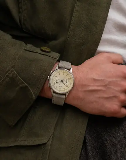 Suede watch strap in grey-beige by vild Hamburg on a vintage Tissot Heritage Chronograph on the wrist of a man wearing a dark green jacket and a white T-shirt.